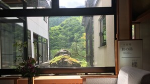 A room with a large window overlooking a garden, a wooden bench, a small table with a vase, and a sign indicating a public bath. - Okada Ryokan Warakutei (Takayama)