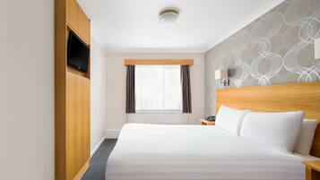 Habitación de hotel con una cama grande, televisor montado en la pared, ventana con cortinas, mesita de noche con lámpara y pared con diseño.