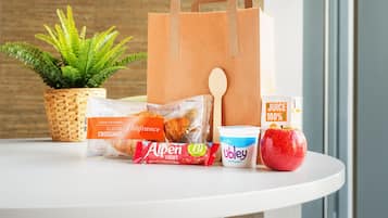Una mesa con una bolsa de papel marrón, una hogaza de pan, un paquete de croissants, un envase de yogur, una manzana roja y un cartón de jugo.