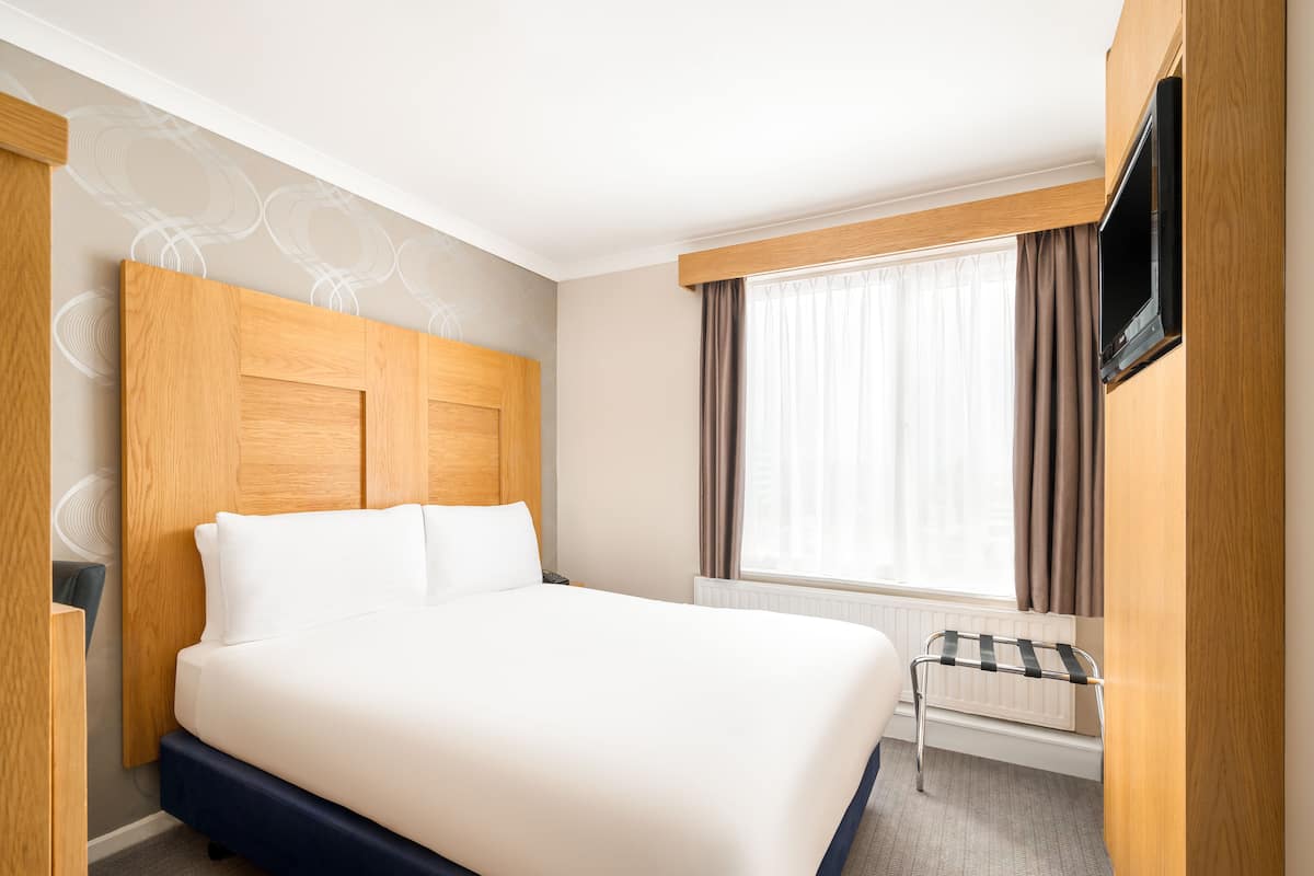 A hotel room with a large bed, a TV mounted on the wall, and a window with curtains.