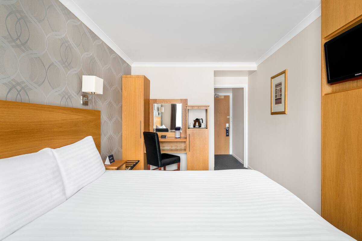 A hotel room with a large bed, a desk with a chair, a mirror, a television, and a framed picture on the wall.