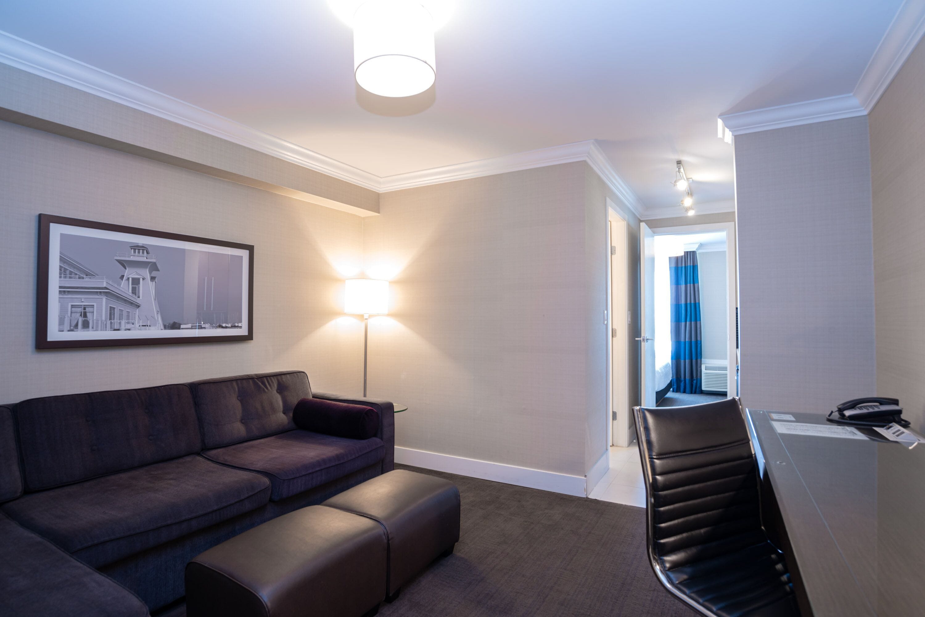 Et moderne hotelværelse med en mørk lædersofa, en gulvlampe, et skrivebord med telefon og et billede i ramme på væggen.
