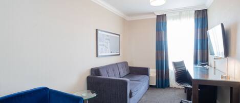 Habitación de hotel con sofá gris, sillón azul, pequeña mesa de vidrio, escritorio con silla y un cuadro en la pared.