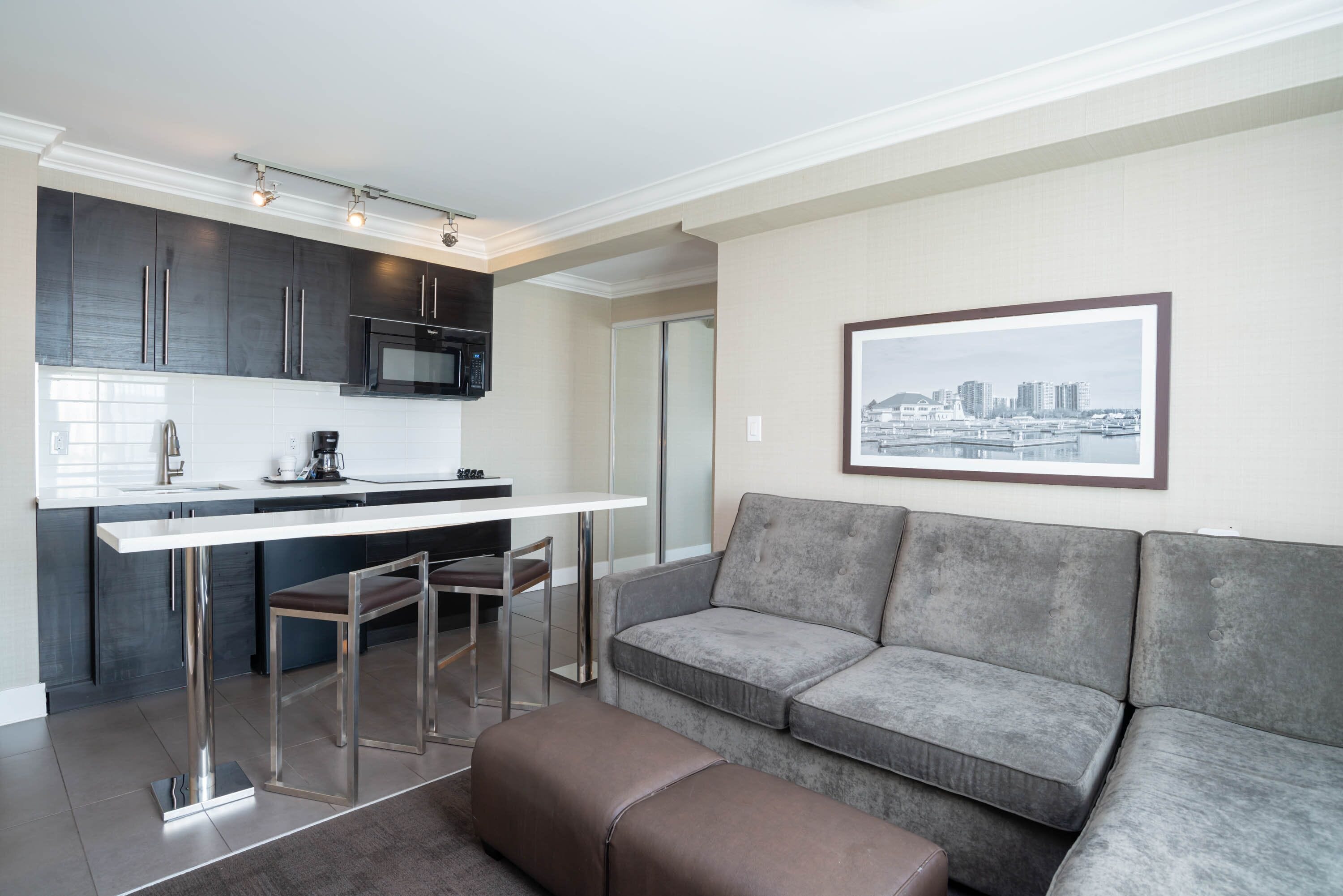 A modern kitchen with dark cabinets, a white countertop, and stainless steel appliances. There is a grey sofa with a framed cityscape picture on the wall.