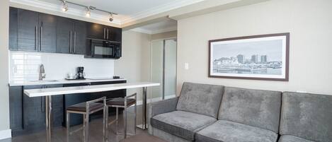 A modern kitchen with dark cabinets, a white countertop, and stainless steel appliances. There is a grey sofa with a framed cityscape picture on the wall.