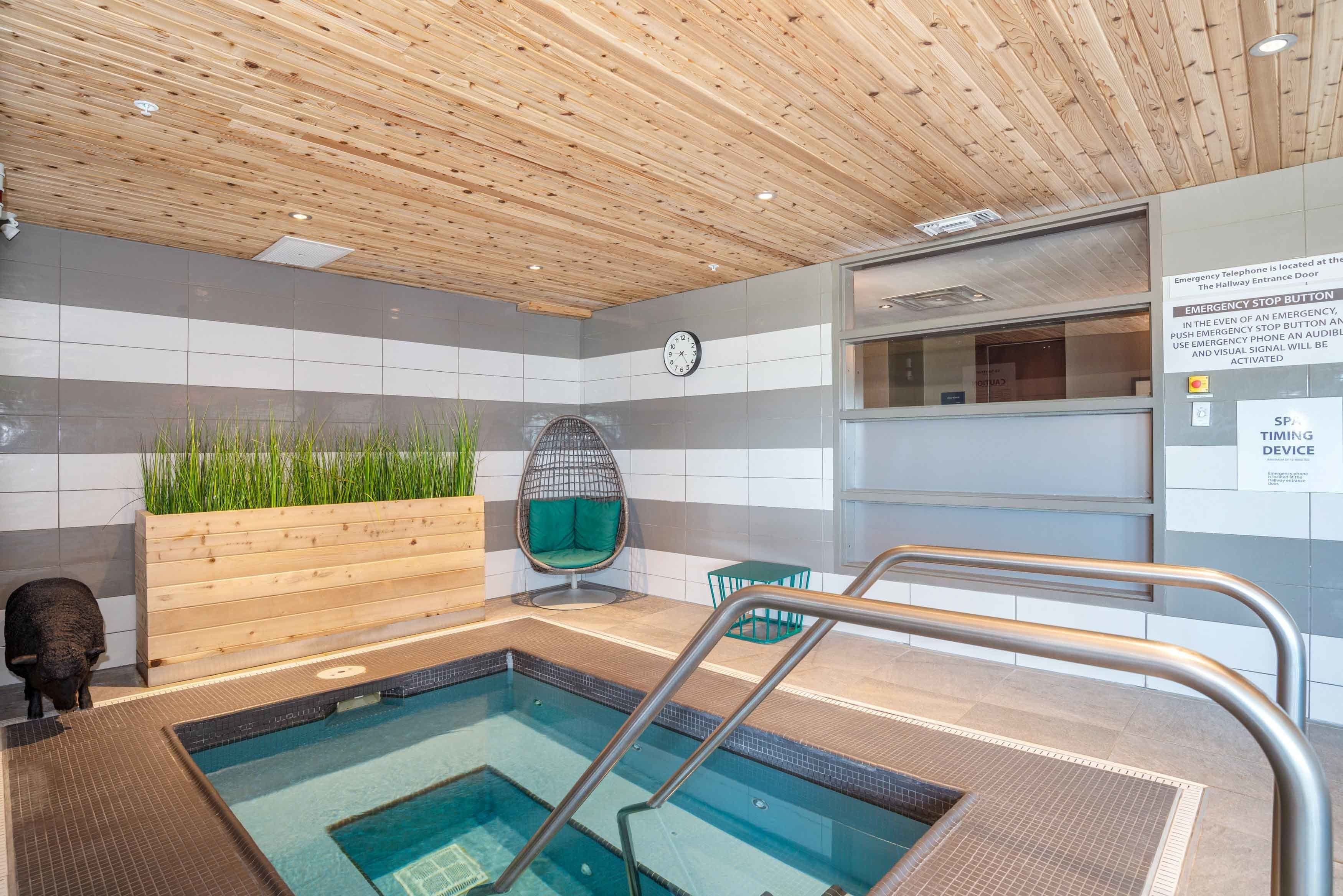 A modern indoor pool area with a wooden ceiling, a clock, a green chair, and a spa device sign.