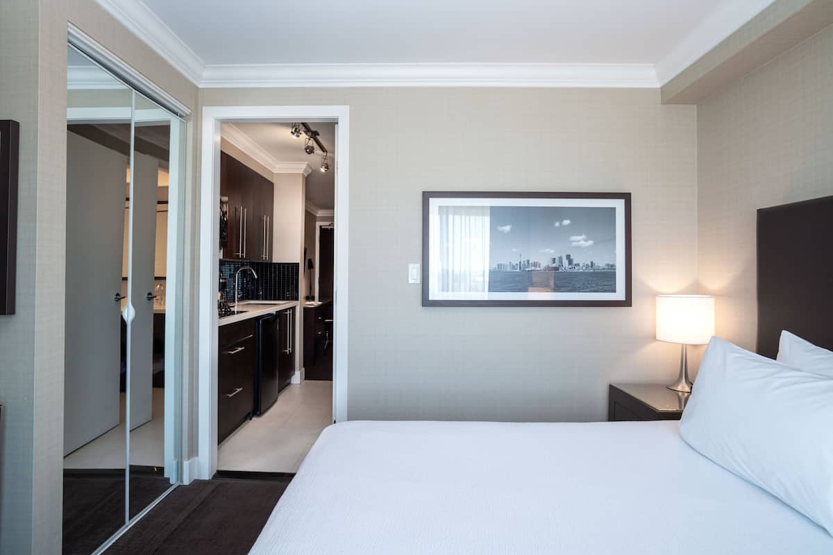 A hotel room with a bed, a nightstand with a lamp, a framed cityscape picture on the wall, a closet, and a kitchen area visible through an open door.