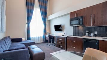 A modern living room with a sofa, ottoman, and glass coffee table. A flat-screen TV is mounted on the wall, and a kitchen area with a microwave, coffee maker, and dishwasher is visible.