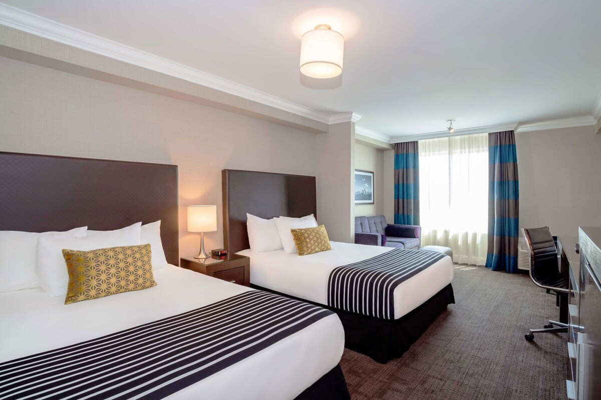 A hotel room with two beds, a desk, a chair, and a view of the city.