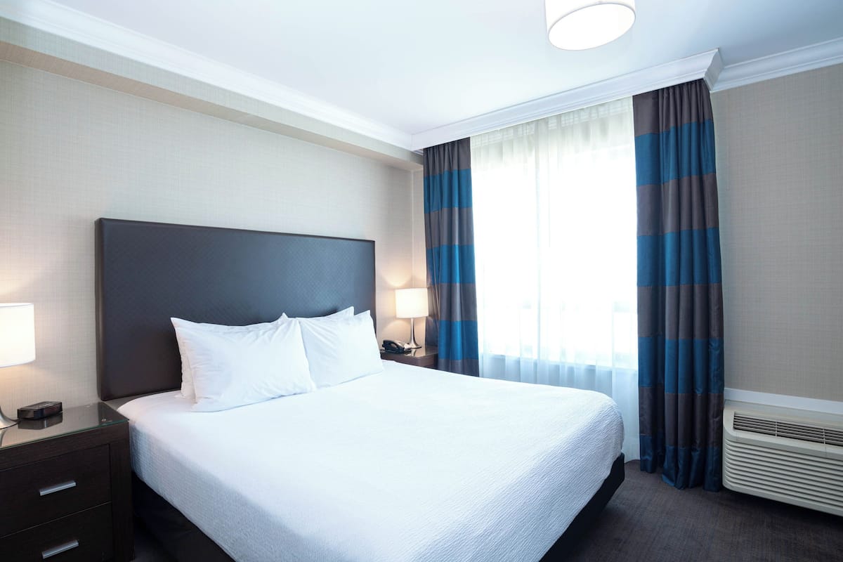 A hotel room with a large bed, a bedside table with a lamp, a nightstand with a phone, and a window with curtains.