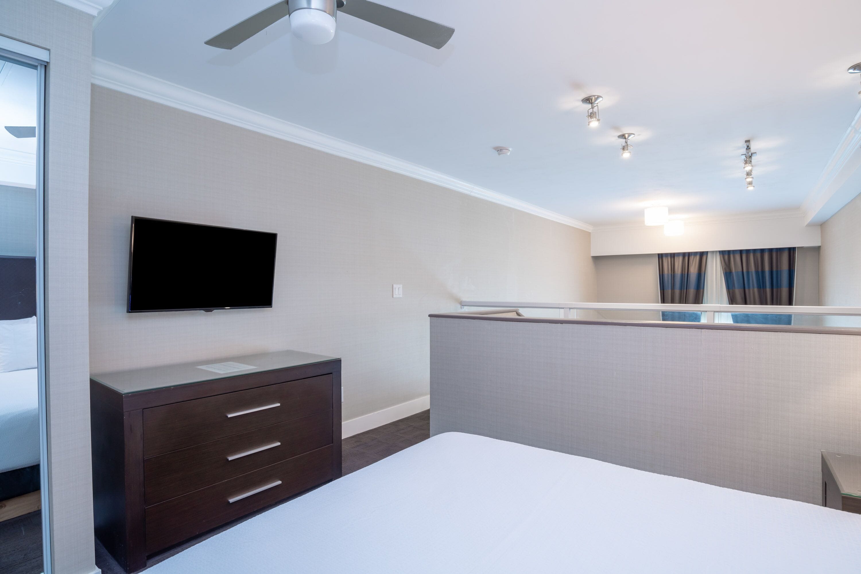 Een hotelkamer met een bed, een aan de muur bevestigde televisie, een dressoir, een plafondventilator en een raam met gordijnen.