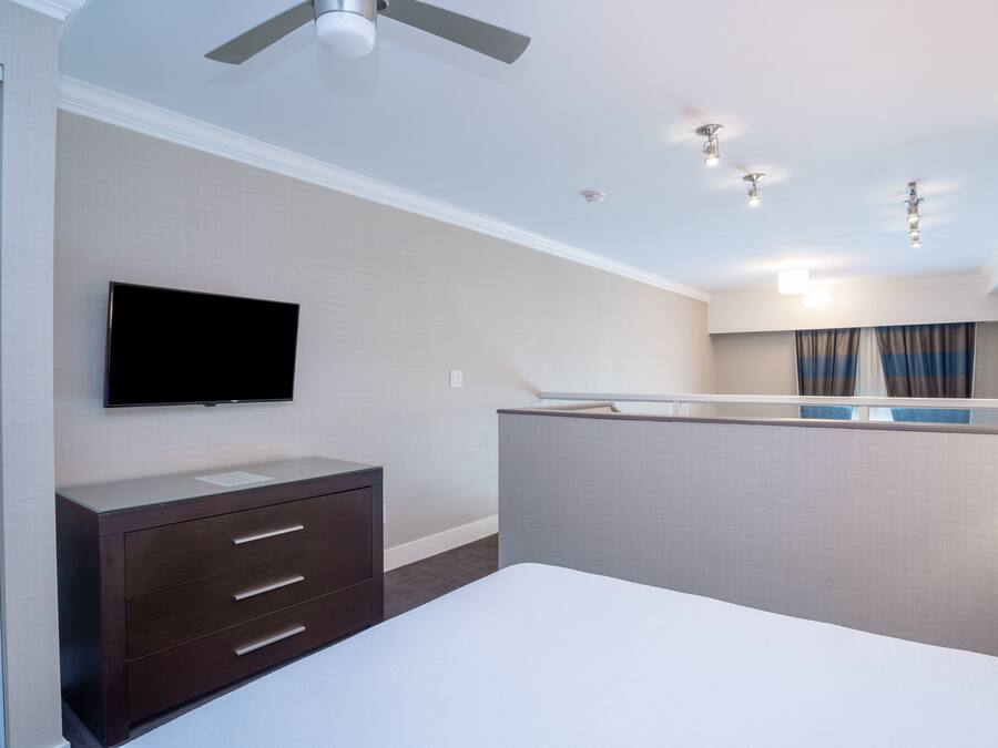 Een hotelkamer met een bed, een aan de muur bevestigde televisie, een dressoir, een plafondventilator en een raam met gordijnen.