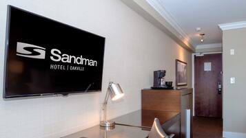 Habitación de hotel con televisión de pantalla plana montada en la pared, escritorio con lámpara, una silla, control remoto y cafetera sobre una encimera.