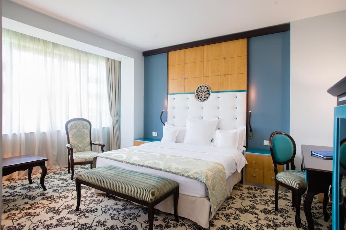 A hotel room with a large bed, two chairs, a bench, a desk, and a nightstand.