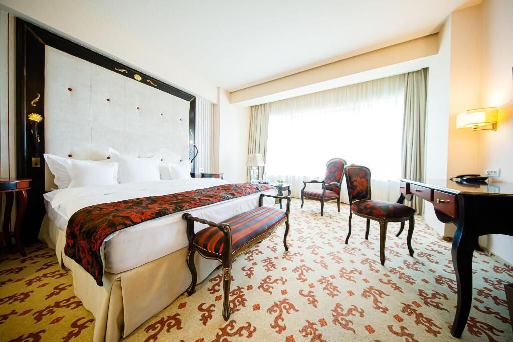 A hotel room with a large bed, a bench, two chairs, a desk, and a window with curtains.