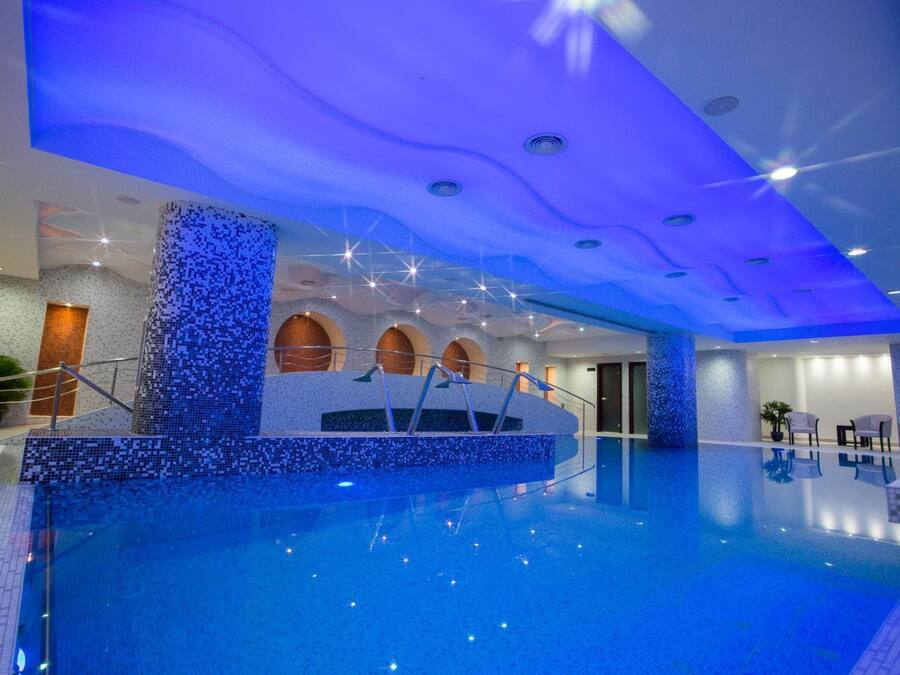 A large indoor swimming pool with a blue mosaic design on the walls and ceiling, a central staircase, and a seating area with chairs.