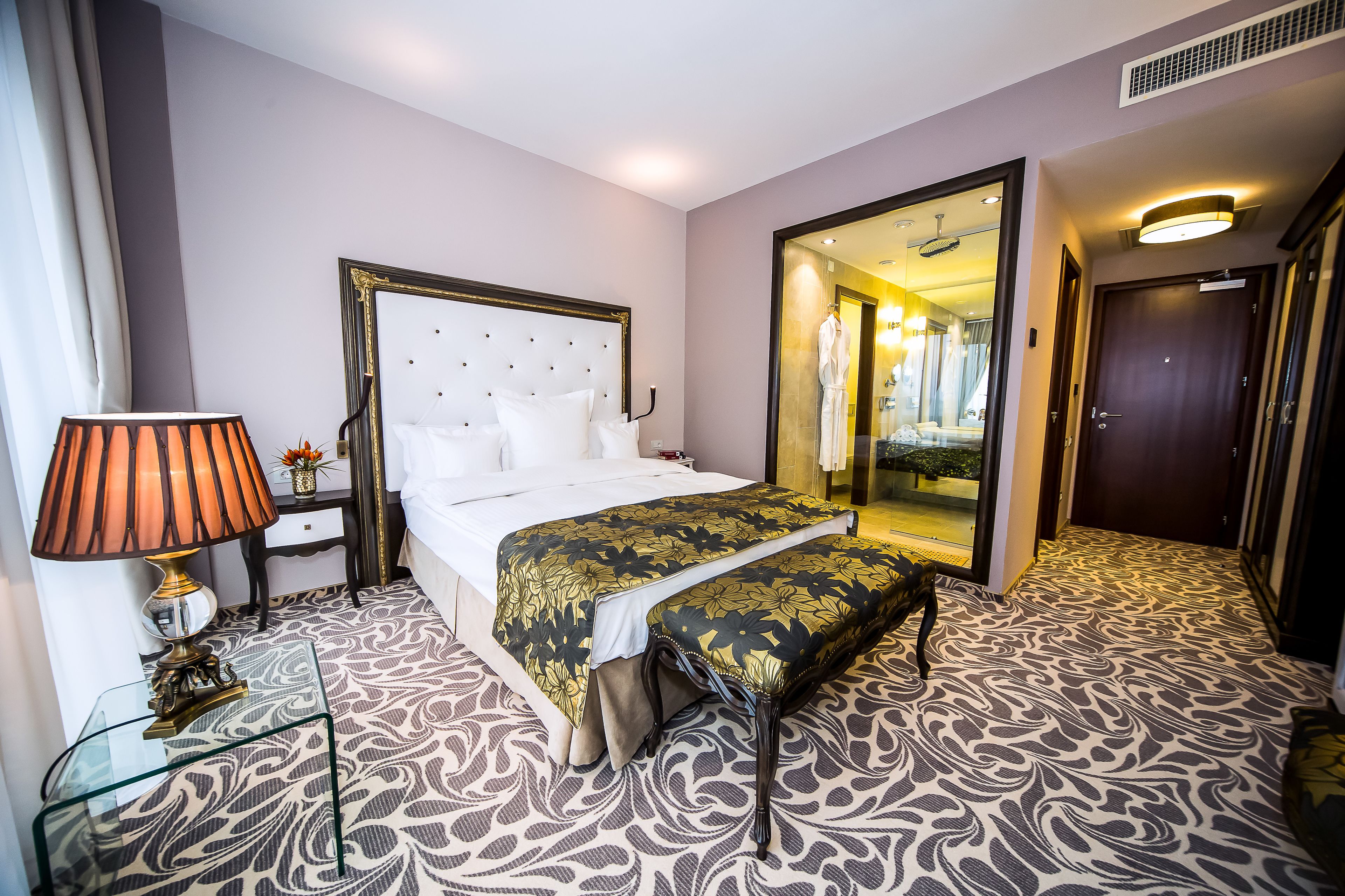 A hotel room with a large bed, a bedside table with a lamp, a bench, and a mirror.