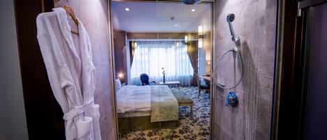 Un baño moderno con cabina de ducha de vidrio, un albornoz blanco colgado en la pared y una cama con cubrecama blanco y una manta verde.