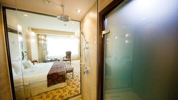 Habitación de hotel con cama, escritorio, silla, zona de ducha y puerta de vidrio.
