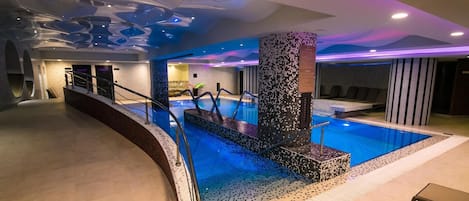 Una piscina interior moderna con un diseño de mosaico único, una zona de estar y una escalera que sube a un nivel superior.
