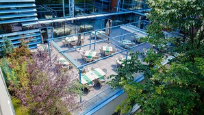 Een terras buiten met tafels en stoelen, omgeven door glazen balustrades en begroeiing.
