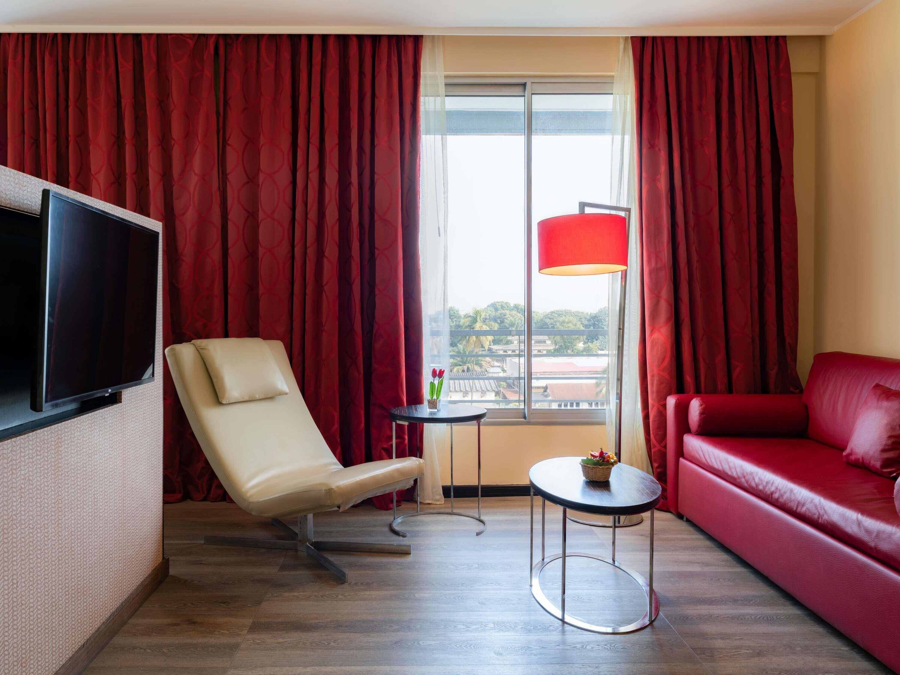 A hotel room with a TV, a red sofa, a red chair, a small table with a lamp, and a window with a view of buildings.