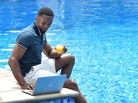 Una persona sentada junto a una piscina con una computadora portátil y un vaso de jugo de naranja.. Pullman Kinshasa Grand Hotel