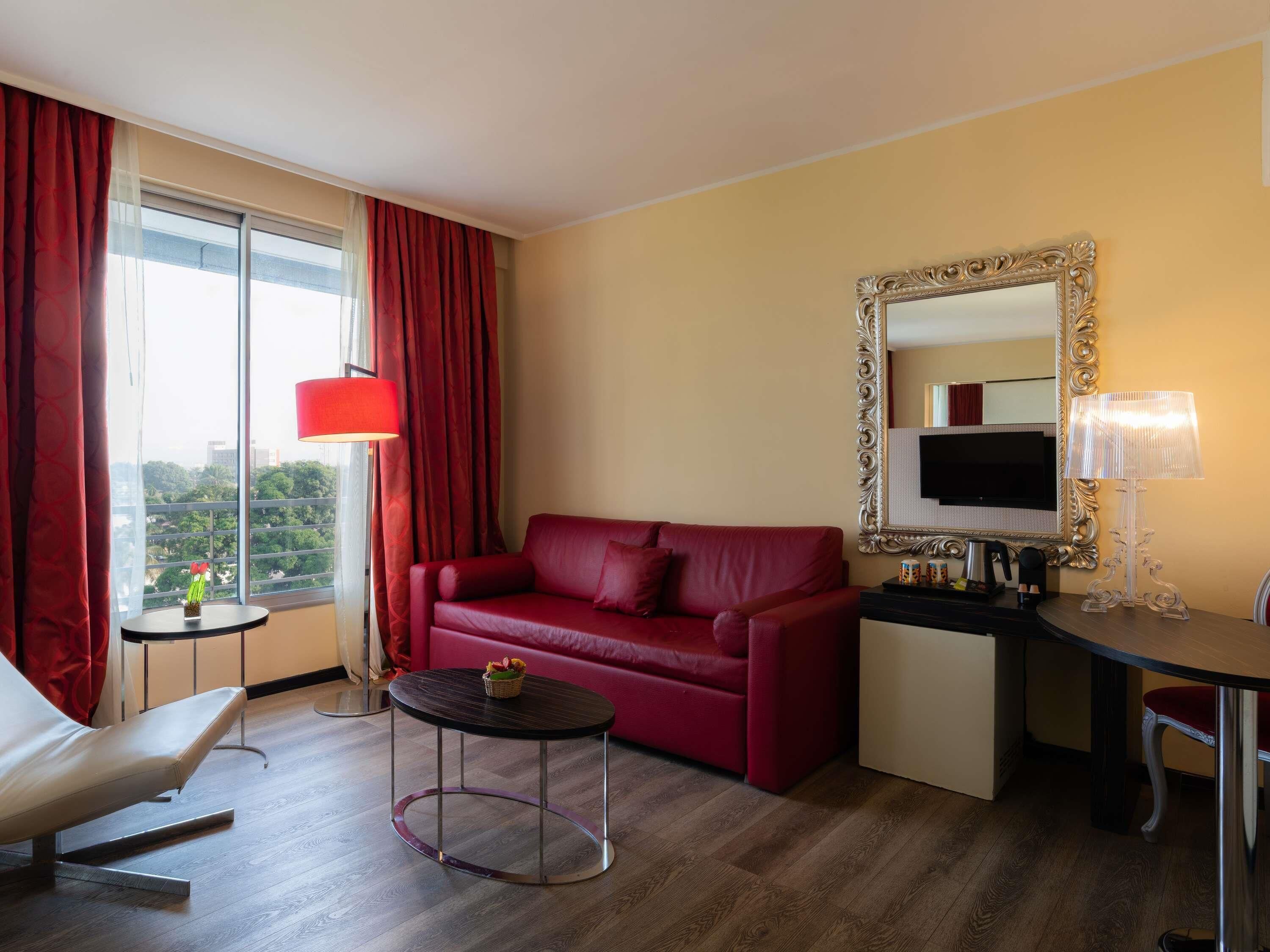 A hotel room with a red sofa, a small round table, a desk with a TV, and a large window with curtains.