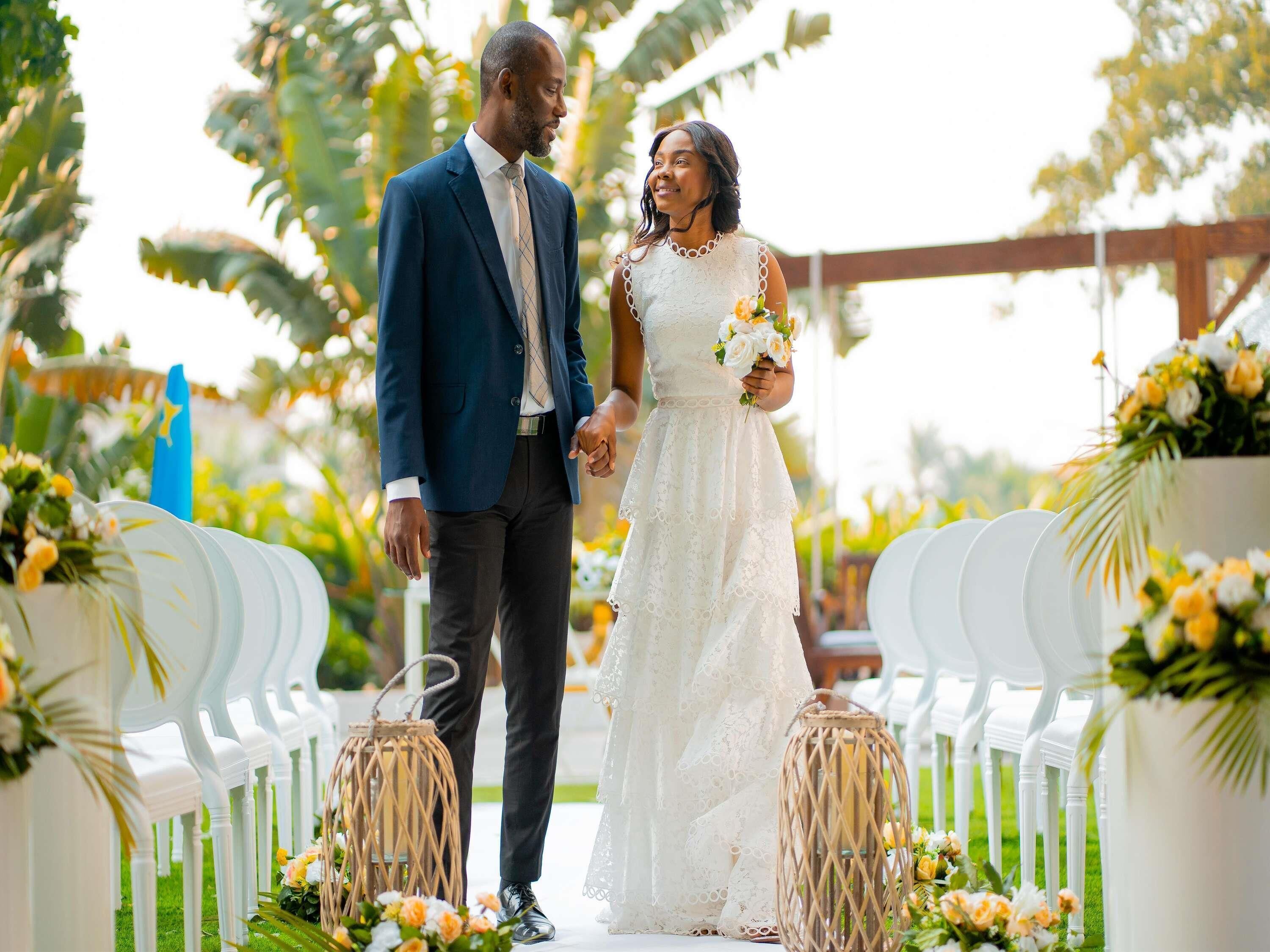 Un couple marchant dans une allée bordée de chaises blanches et illuminée de lanternes en osier, le tout entouré de verdure.