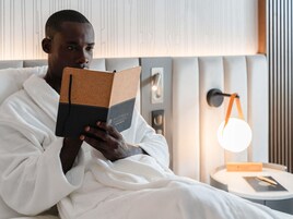 Uma pessoa vestindo um roupão branco lendo um livro em um quarto com uma cama, uma mesa de cabeceira com abajur e uma parede com linhas verticais.