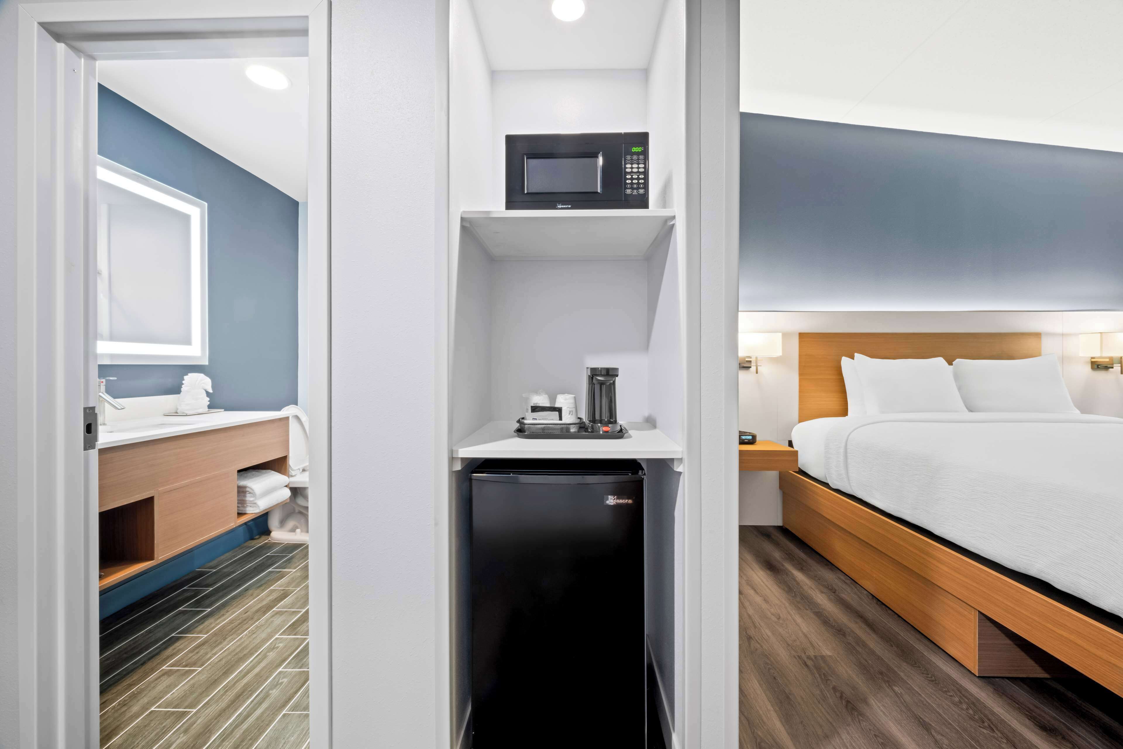 Quarto de hotel compacto com cama, micro-ondas, mini-geladeira, pia e espelho.