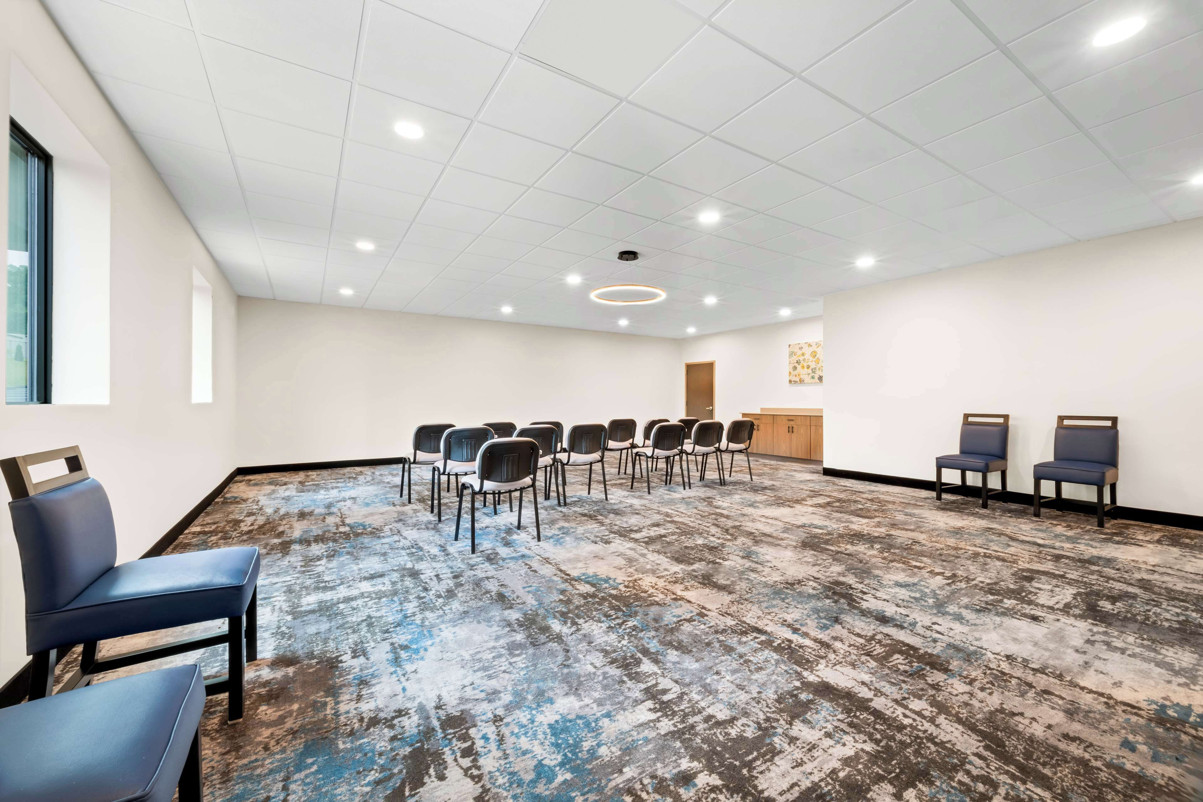 A conference room with a white ceiling, recessed lighting, a patterned carpet, and rows of chairs.