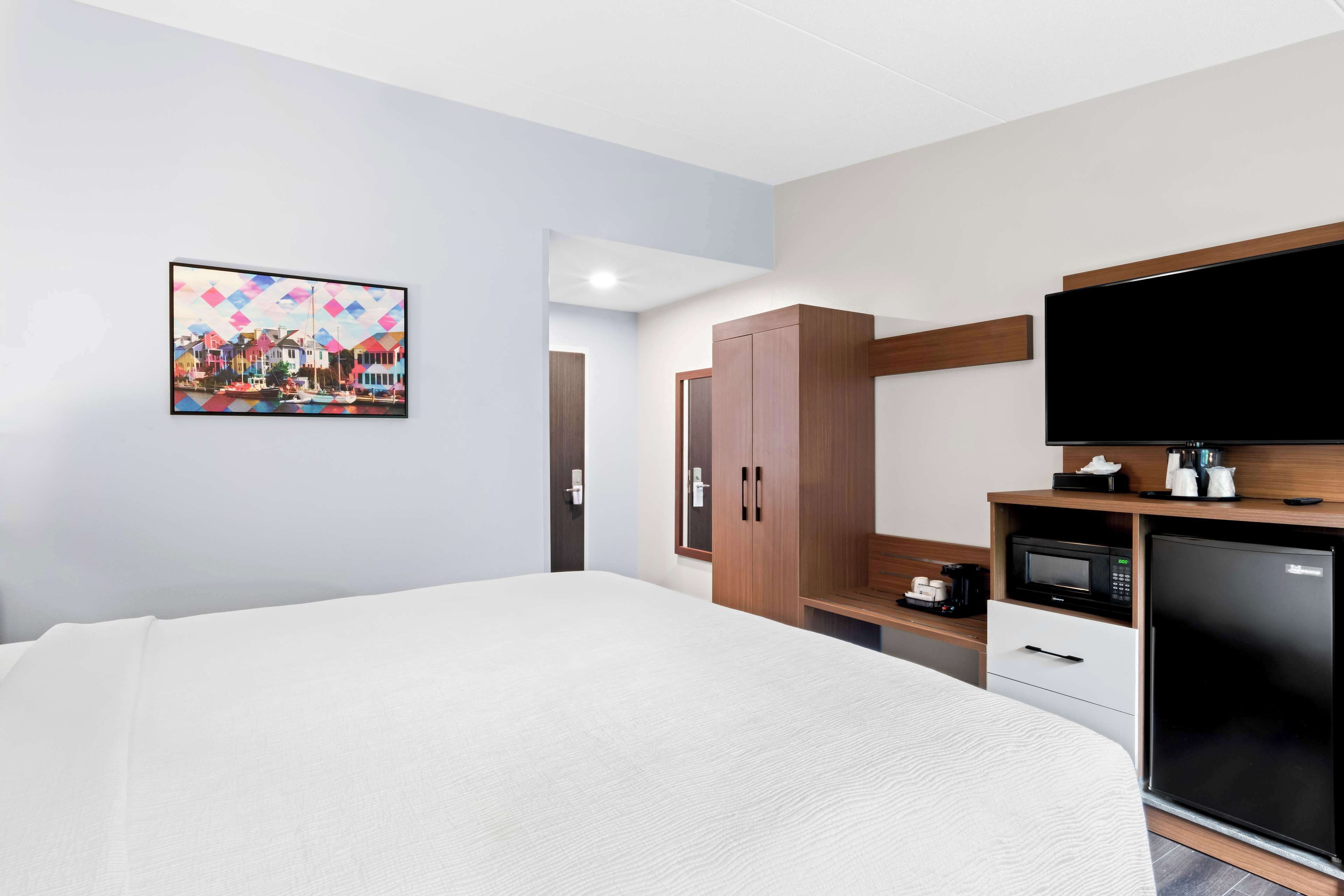 Una habitación de hotel con una cama grande, un televisor de pantalla plana, un microondas, un refrigerador, un armario de madera y un cuadro en la pared.