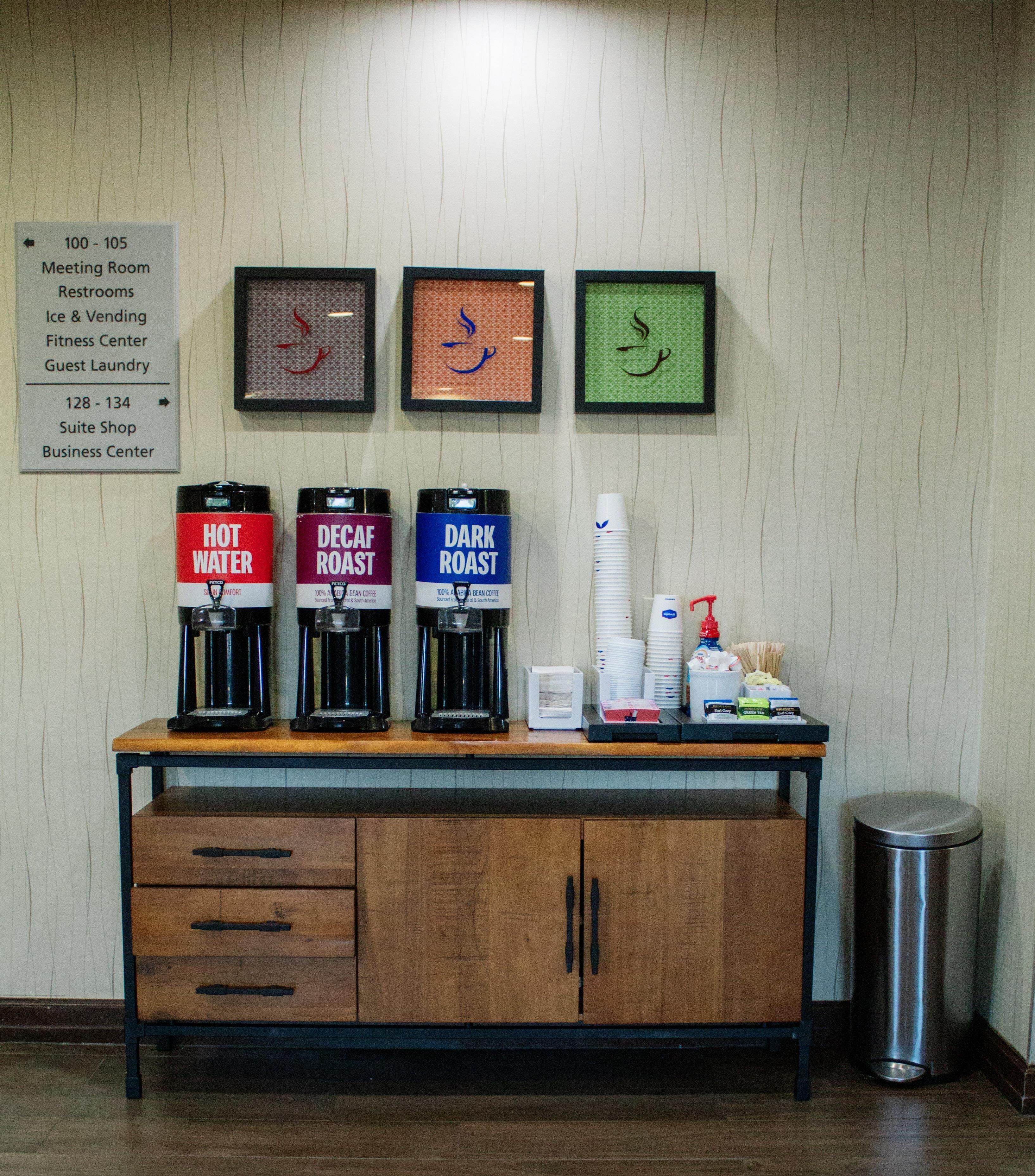 Un espace dédié au café, équipé de trois machines à café étiquetées « EAU CHAUDE », « CAFÉ DÉCAFÉINÉ » et « CAFÉ TORRÉFIÉ », ainsi qu’un panneau indiquant les numéros des chambres et les équipements disponibles.
