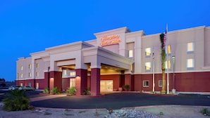 Un hotel con un llamativo letrero de 'Hampton Inn & Suites', un estacionamiento y un asta con la bandera estadounidense.