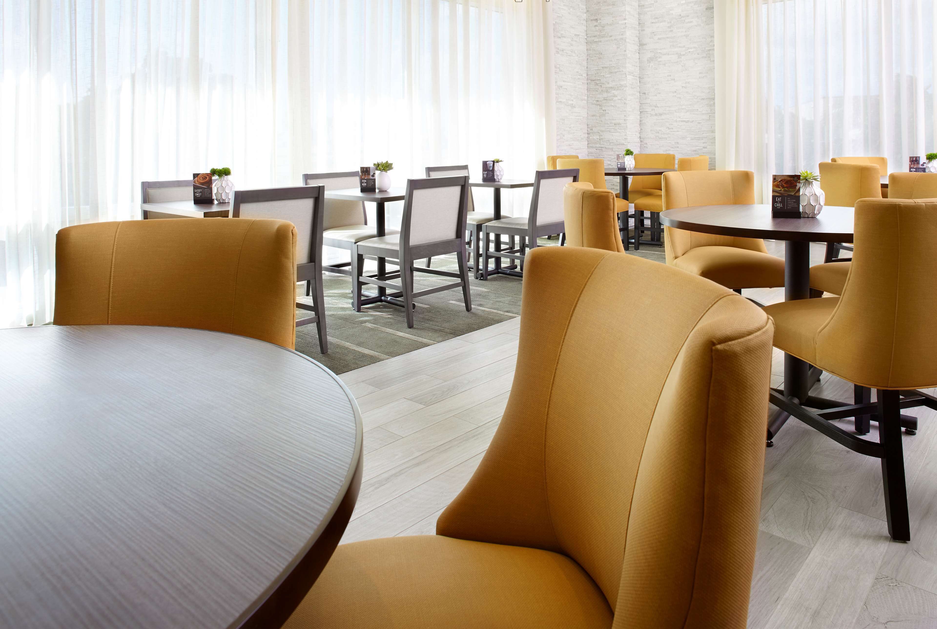 A modern dining area with round tables and yellow chairs, featuring a light-colored wood floor and large windows with sheer curtains.