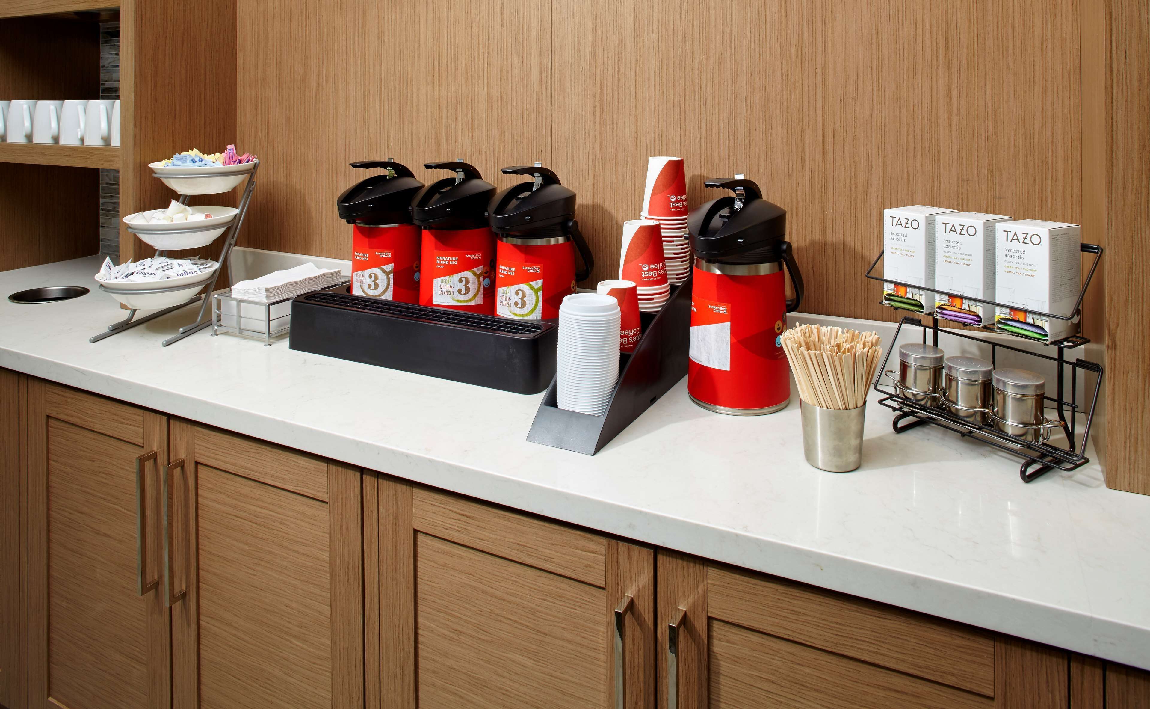 Zona de recepción de un hotel con mostrador, estación de café y diversas comodidades como tazas, platos y recipientes de comida.