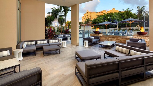 A modern outdoor lounge area with sofas, tables, and a fire pit, overlooking a pool and palm trees.