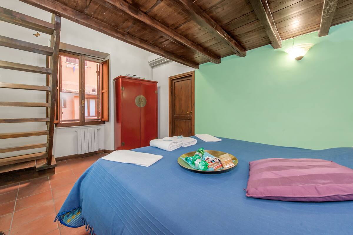 Una habitación con una cama, una escalera de madera, una puerta roja y una ventana.