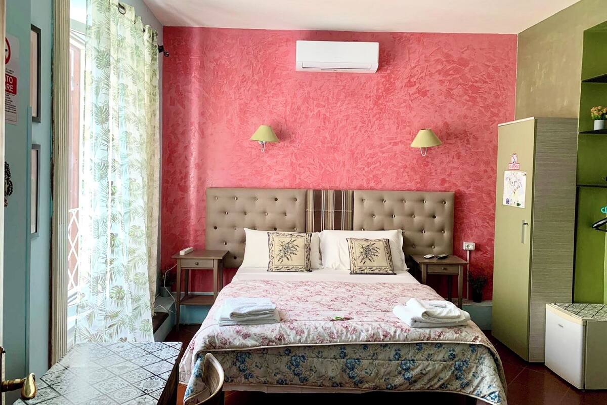 Habitación de hotel con pared rosa texturizada, cabecera capitonada, dos lámparas de noche, cama con ropa de cama floral y un refrigerador pequeño.