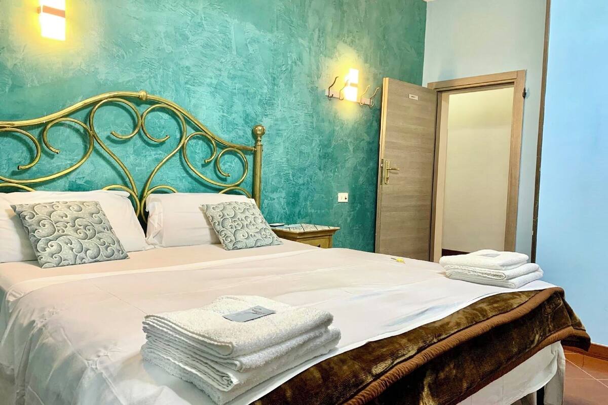 Una cama bien hecha con sábanas blancas y una manta marrón, una mesita de noche con una lámpara, y una pared color turquesa con un accesorio de iluminación.