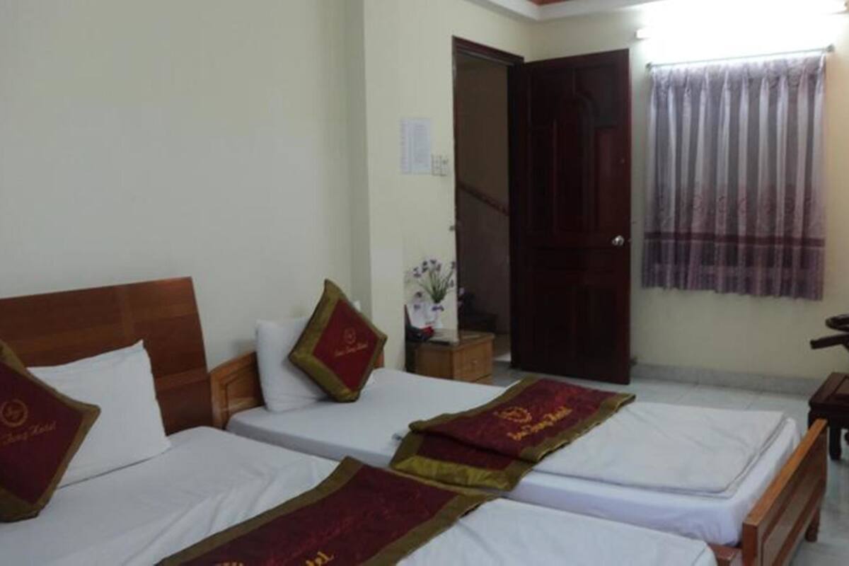 A hotel room with two beds, a wooden headboard, a nightstand, a window with curtains, and a door.