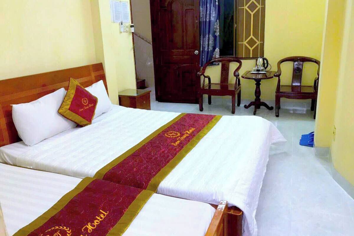 A hotel room with two beds, a wooden headboard, a small table with chairs, and a window with curtains.