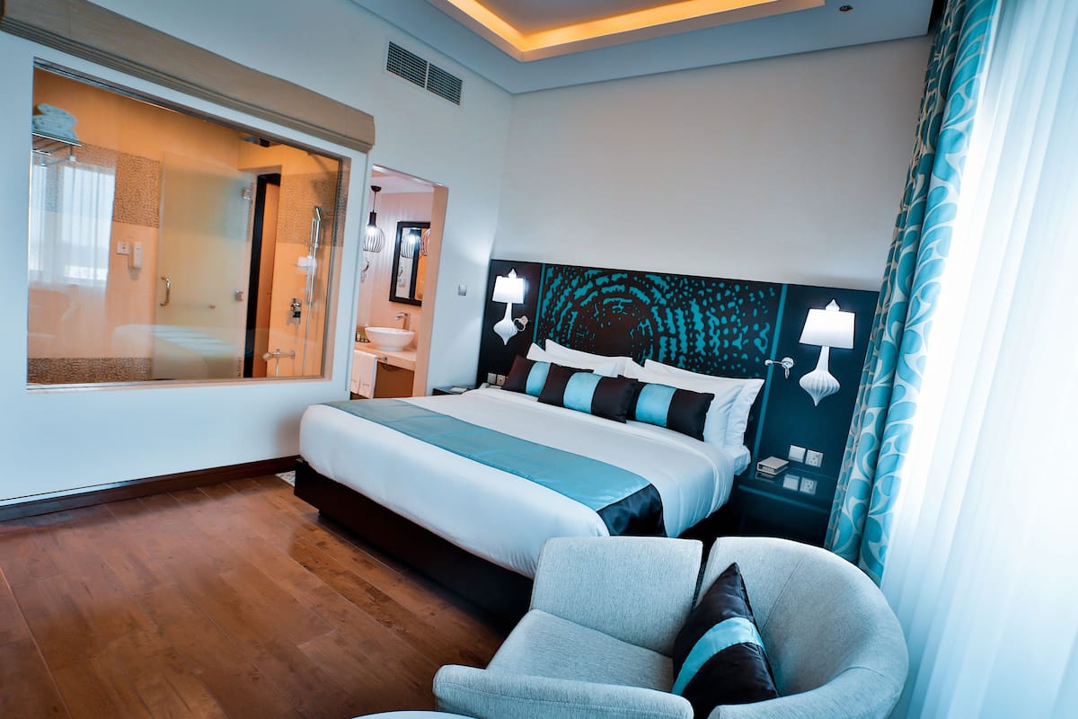 A modern hotel room with a large bed, a seating area, a bathroom with a glass shower enclosure, and a view of the city.