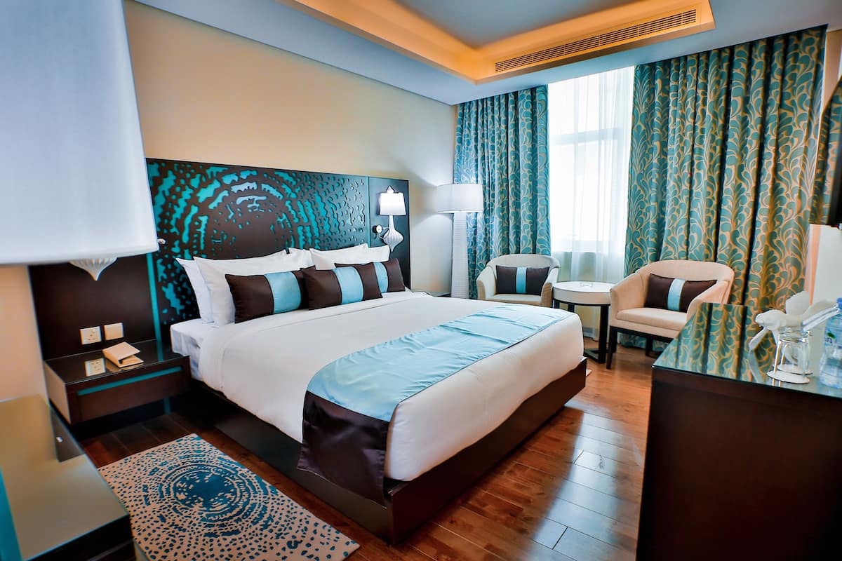 A hotel room with a large bed, a desk, a chair, a lamp, and a patterned rug.