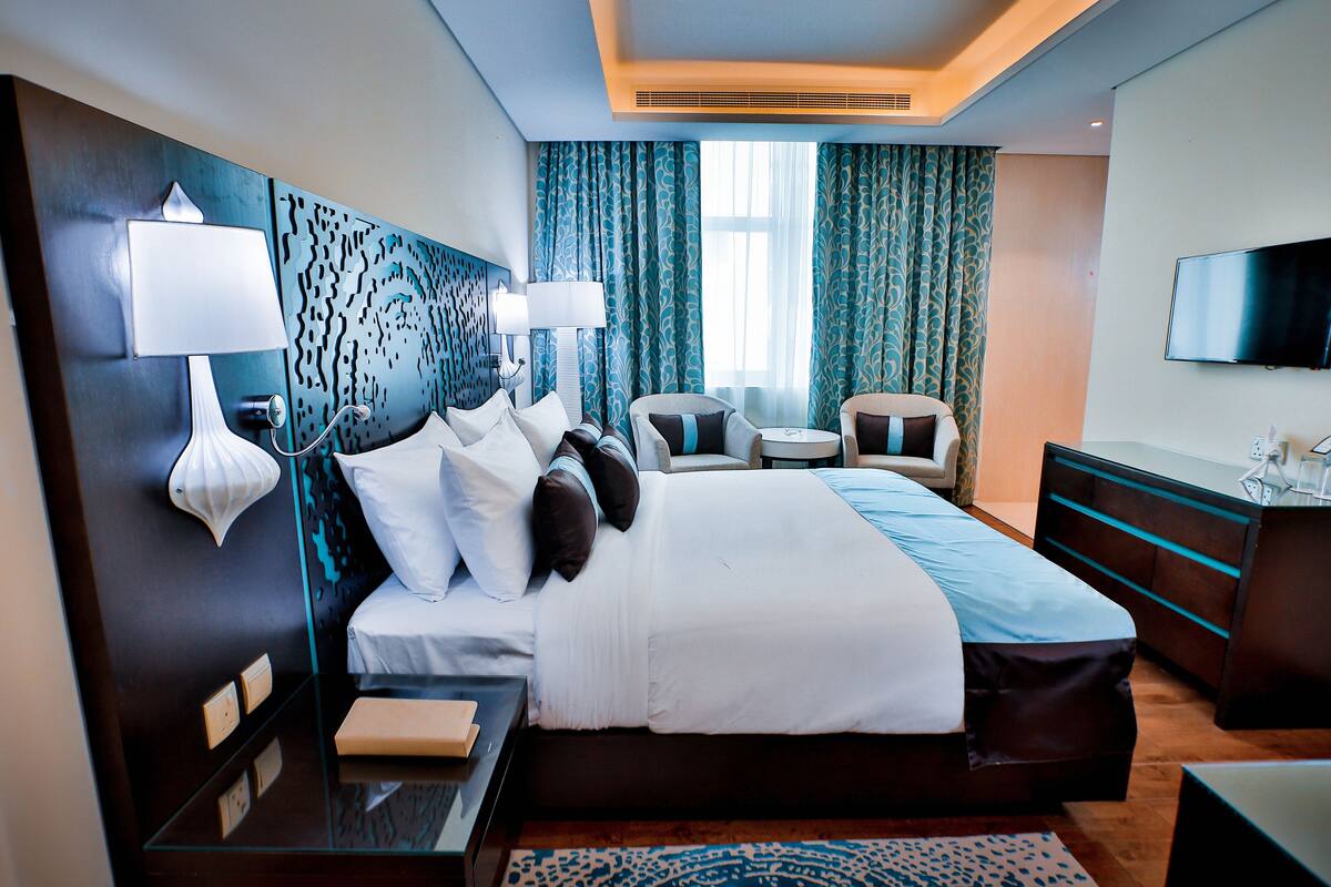 A modern hotel room with a large bed, bedside tables, a desk, a chair, a TV, and a window with curtains.