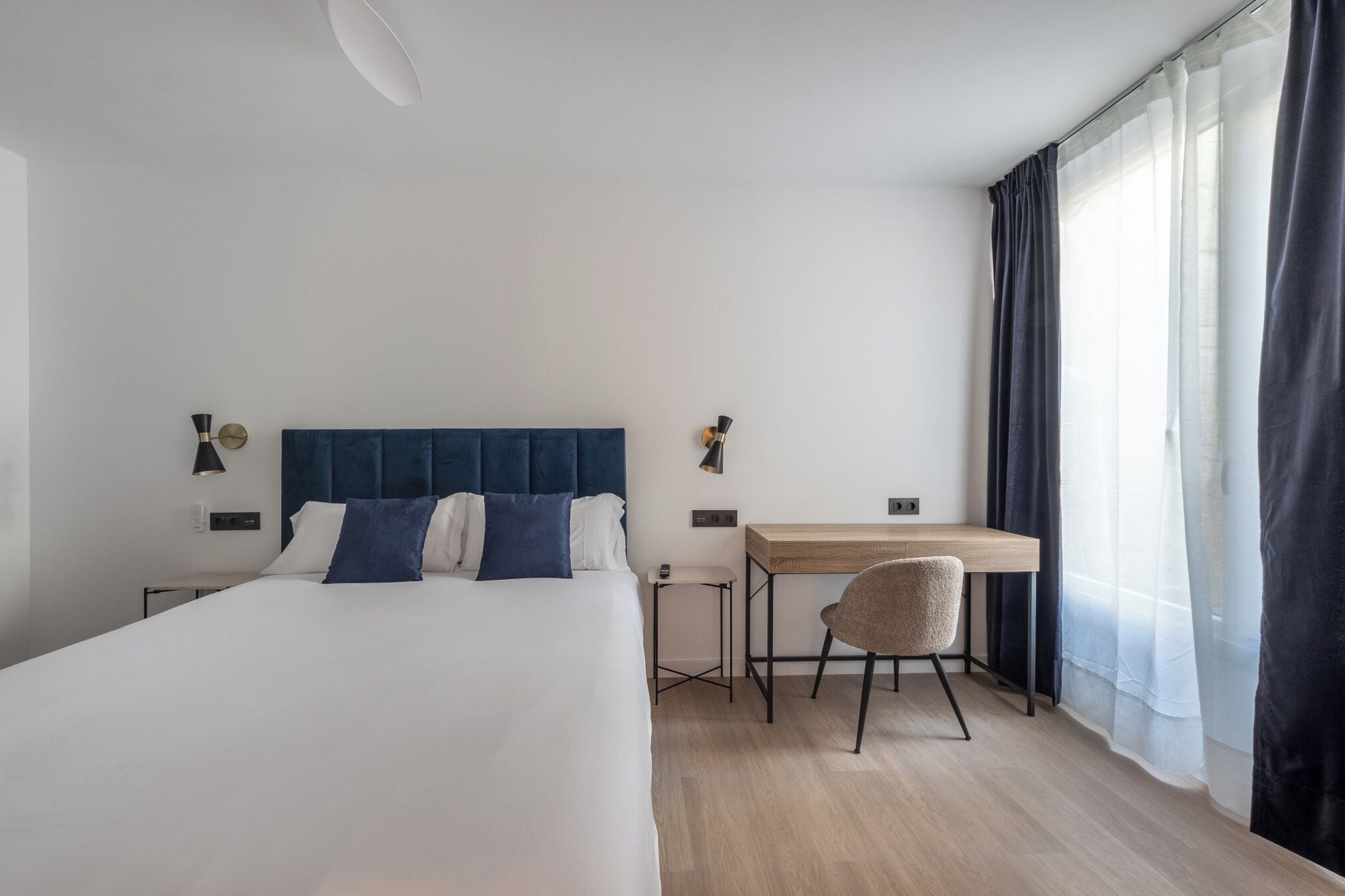 Habitación de hotel con una cama grande, un escritorio y una silla. Hay dos lámparas de pared y una ventana con cortinas.