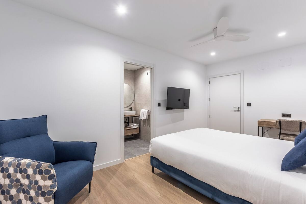 A modern hotel room with a large bed, a blue armchair, a small table, a TV mounted on the wall, and a ceiling fan.