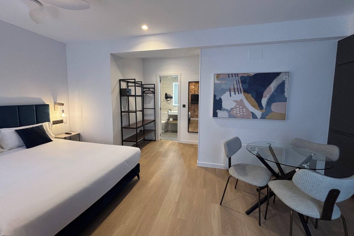 A modern hotel room with a large bed, a desk with chairs, a bookshelf, and a bathroom visible through an open door.