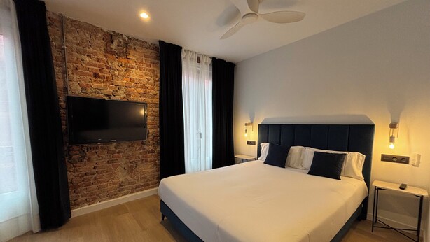 A modern hotel room with a large bed, a flat-screen TV mounted on a brick wall, a ceiling fan, and two bedside lamps. -  Arenal Rooms Bilbao (Bilbao)